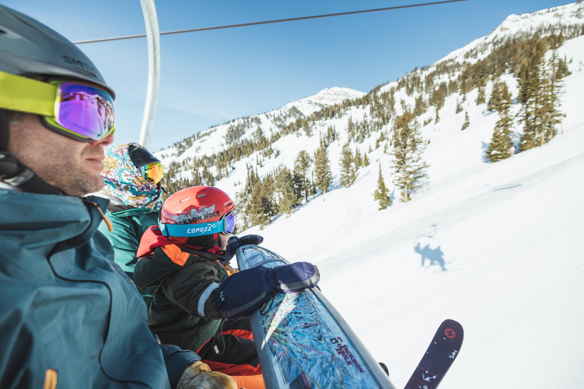 Three skiers in colorful gear ride a snowy mountain chairlift under a clear blue sky.